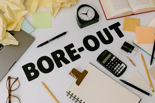 Top view van een rommelig bureau met het woord “BORE-OUT” in grote zwarte letters, omringd door een klok, papieren, kantoorartikelen en een open boek.