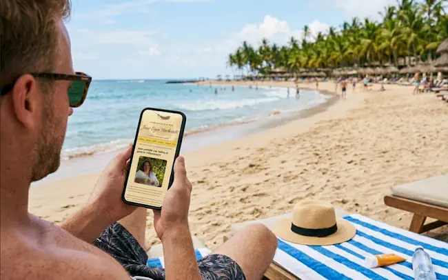 Man op het strand die moe tijdens zijn vakantie op zijn telefoon de blog van Mieke Loor, Jouw Eigen Veerkracht, leest