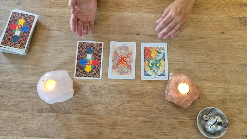 Tarotkaartlegging met drie kaarten op een houten tafel, geflankeerd door twee brandende theelichtjes in zoutlampen en handen die het legpatroon toelichten.