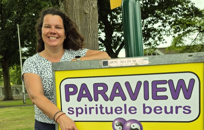 Mieke Loor van praktijk Jouw Eigen Veerkracht glimlacht naast een geel bord met paarse tekst "Paraview spirituele beurs" op een buitenterrein.