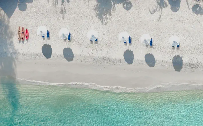 wit-strand-blauw-water-zomer-parasol Luchtfoto van een leeg strand met witte parasols, ligbedden en kajaks langs de turquoise zee.