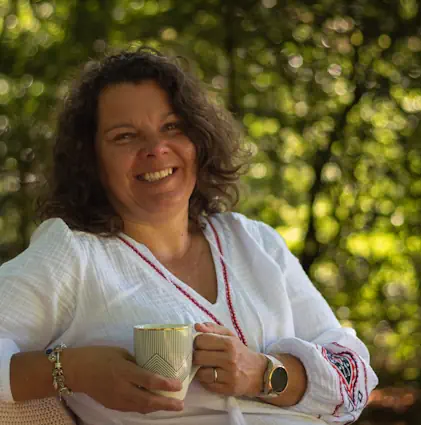 Mieke Loor zit ontspannen buiten met een kop thee, warm glimlachend in een groene omgeving