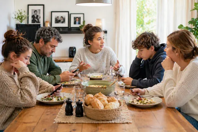 Gezin aan tafel waarbij één persoon wil praten maar anderen afgeleid zijn – moeite met praten over gevoelens – therapie Mieke Loor