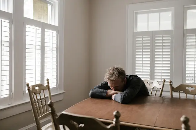 Man ligt uitgeput met hoofd op keukentafel, alleen in lege kamer, depressief na ontslag
